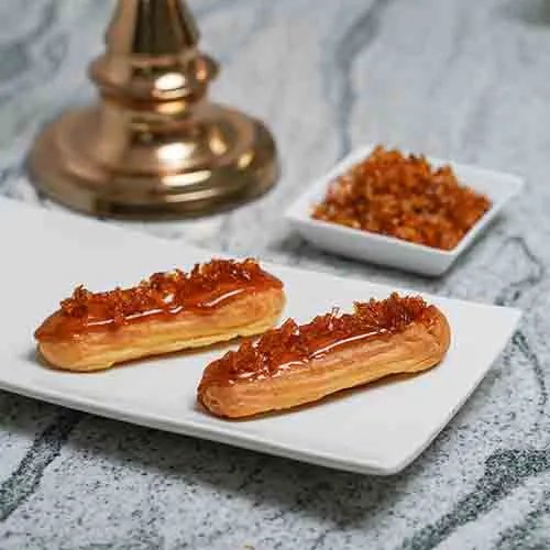 Two eclairs with a caramel crunch topping on a white plate, a dish of the crunch and a brass candlestick behind