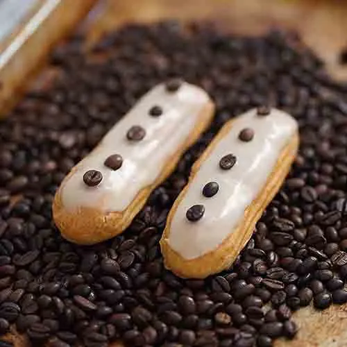 Two coffee-glazed eclairs dotted with chocolate, on a bed of coffee beans