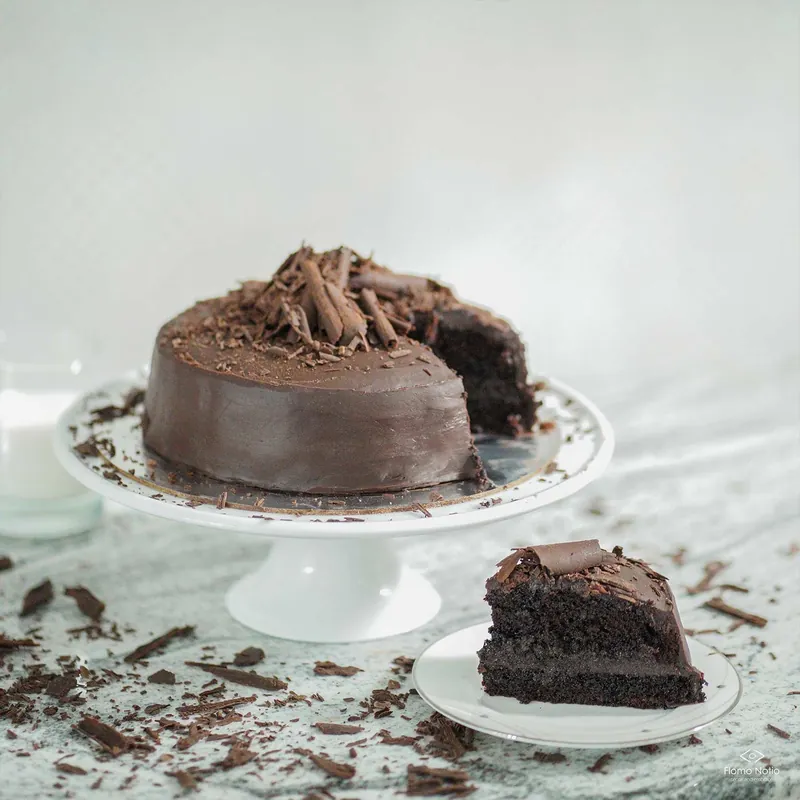 Chocolate cake on a stand with a slice cut out, the slice on a plate in front and shavings scattered on marble
