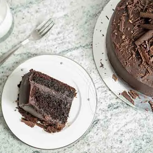 A slice of layered chocolate cake on a white plate with a fork, the rest of the cake beside it on marble
